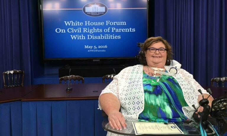 Carrie Ann Lucas, a woman using a wheelchair, sits in front of a screen reading "White House Forum on Civil Rights of Parents with Disabilities."