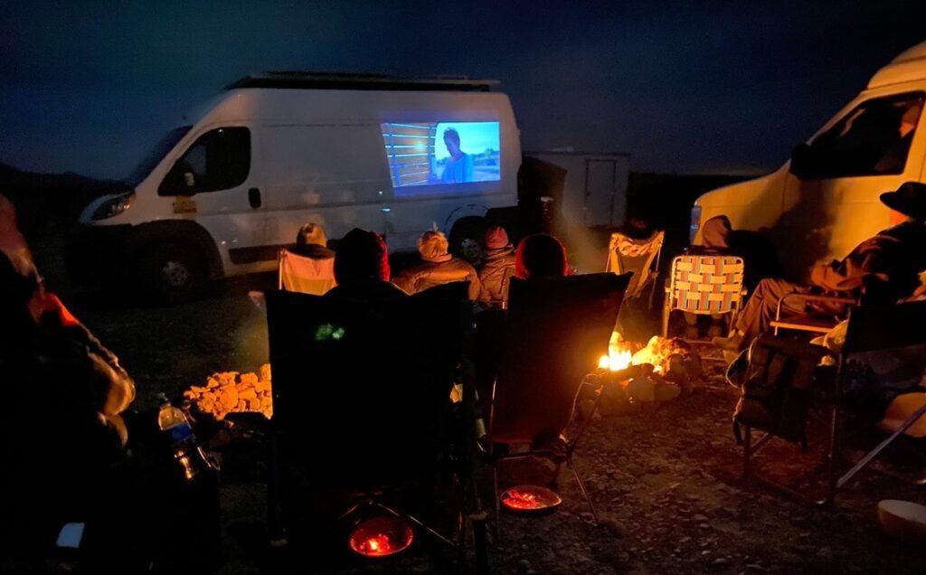 Women gathered around a desert campfire watching a video on the side of a white van
