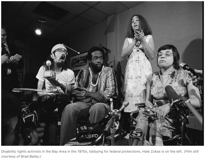 Three people sit in wheelchairs - one white man using mouth navigation, a black man, and a white woman. A woman stands behind them doing ASL interpretation.