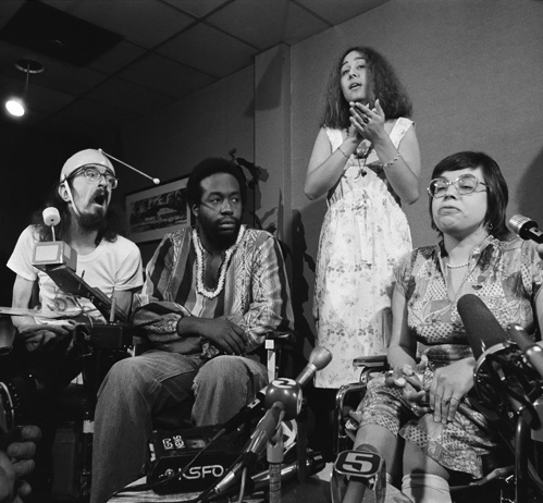 Three people using wheelchairs - a white man using a mouth stick, a Black man, and a white woman. A sign language interpreter stands behind them.