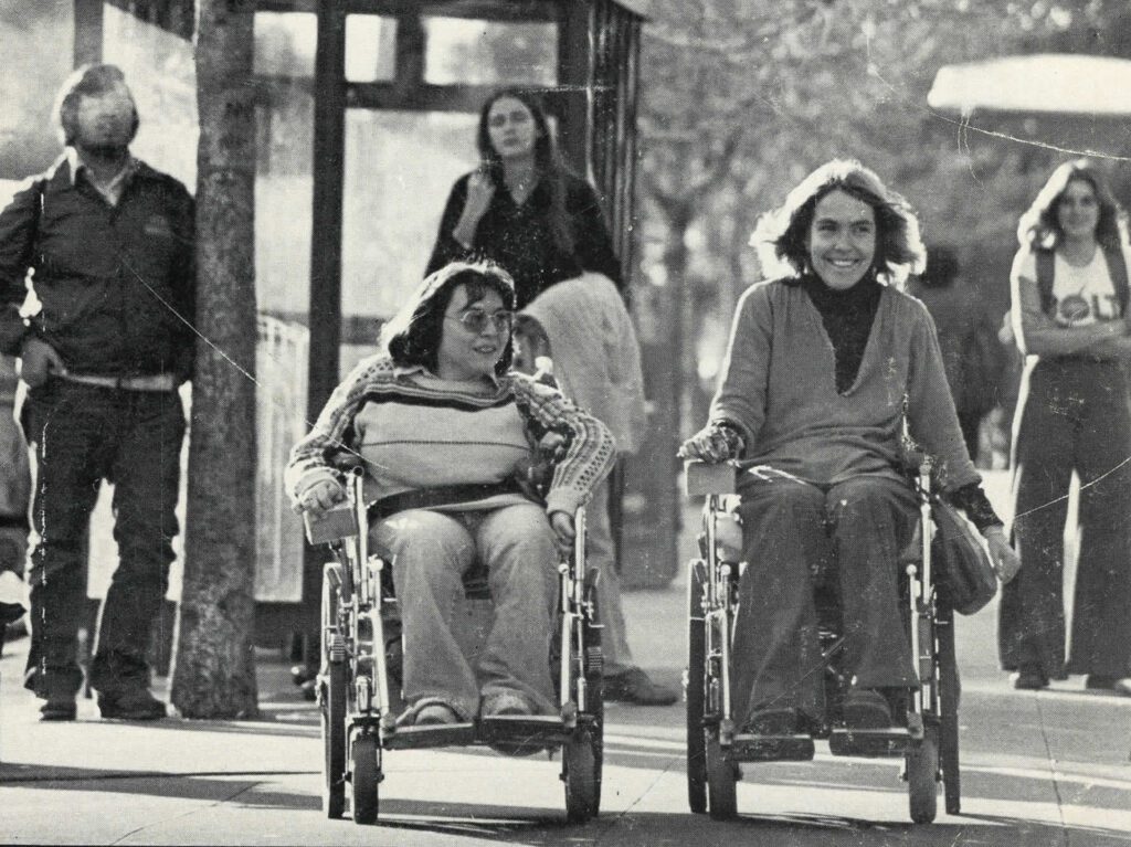 Two women in wheelchairs roll down a city sidewalk.