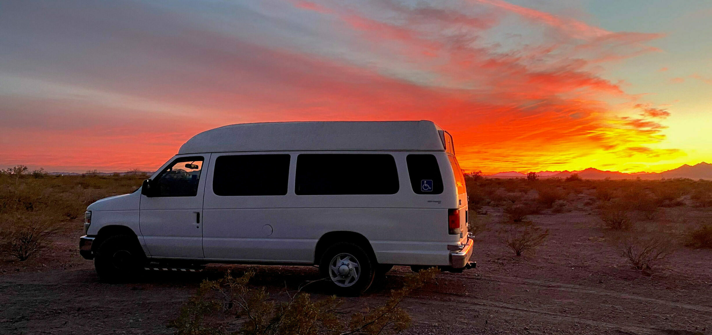 A white van parked in a dirt field with a red, gray, and yellow sky behind