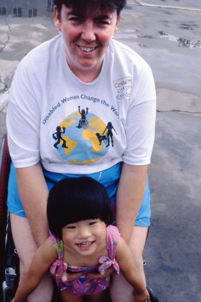 A white woman sits in a wheelchair holding a Asian toddler between her legs.