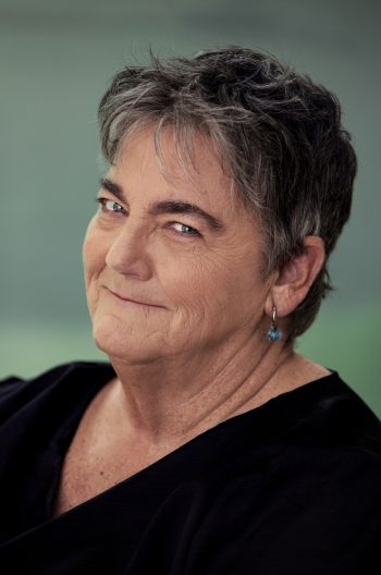 Headshot: Corbett O’Toole – a smiling Caucasian woman in her 60’s with short hair wearing a black v-neck shirt.